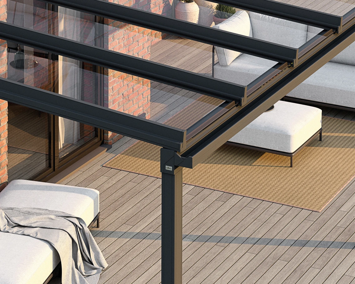 Outdoor patio under a glass canopy with a dark metal frame, wooden deck, brick wall, and cushioned seating on beige rugs.